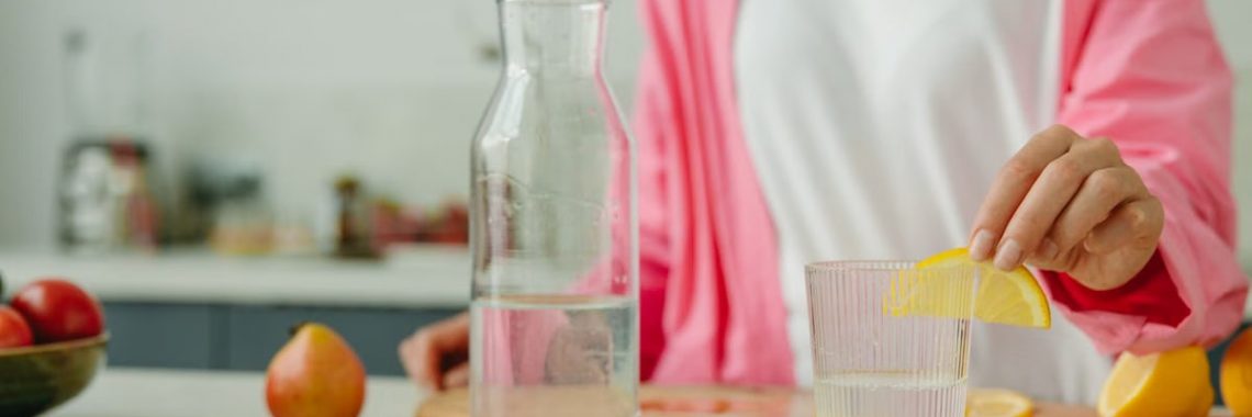 A woman in a pink outfit prepares lemon water in a bright, modern kitchen setting.